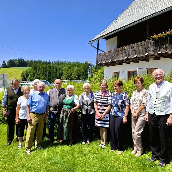 Eine Gruppe älterer Personen in traditioneller Kleidung, die in einem Garten vor einem rustikalen Haus mit Balkon steht.