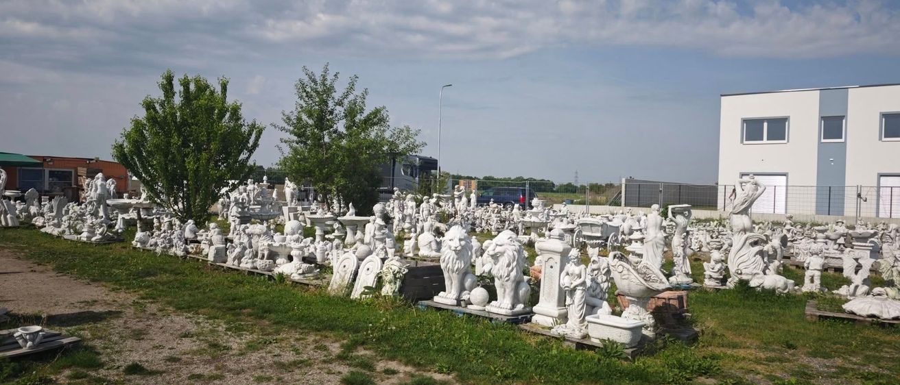 Ein Feld mit vielen weißen Skulpturen, darunter Löwen und Engel, unter einem teilweise bewölkten Himmel. Im Hintergrund befinden sich Bäume und ein Gebäude.