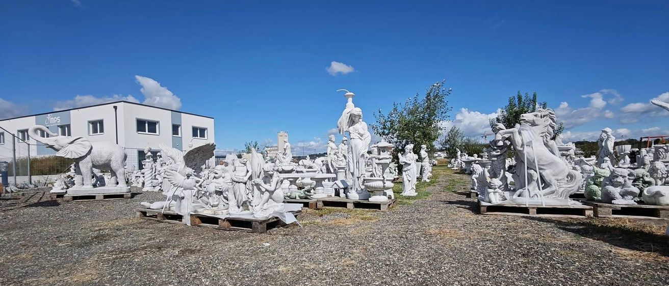 Ein Kiesfeld mit weißen Statuen von Menschen, Tieren und mythischen Kreaturen auf Holzpaletten unter einem blauen Himmel mit Wolken. Ein weißes Gebäude mit mehreren Fenstern ist im Hintergrund sichtbar.