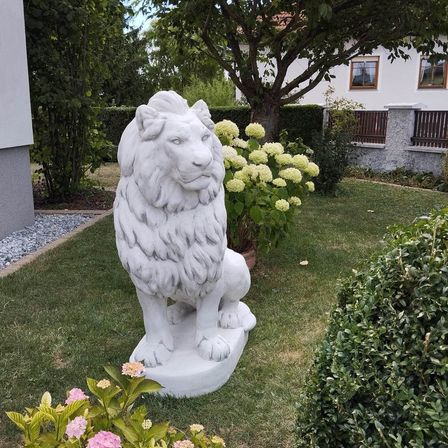 Eine weiße Löwenstatue steht in einem Garten mit rosa Blumen, einem Baum und einem weißen Haus.