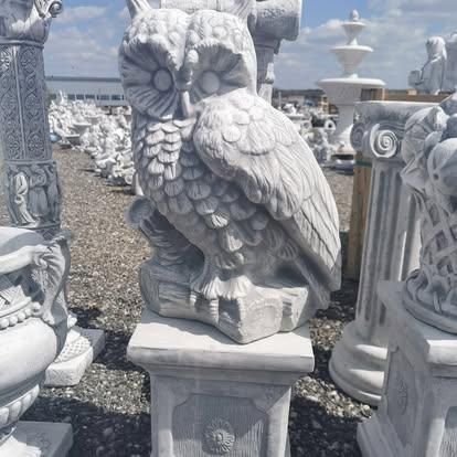 Eine große weiße Marmorstatue einer Eule, die auf einem Steinsockel in einem Garten steht, umgeben von anderen Marmorskulpturen.