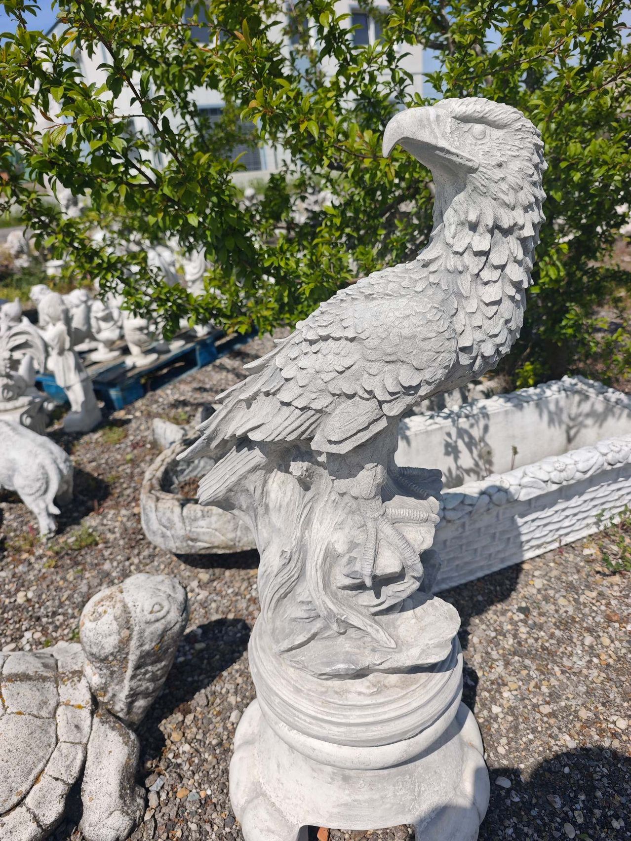 Eine weiße Steinadlerstatue steht prominent in einem Garten, mit detaillierten Federn und einem scharfen Schnabel, umgeben von anderen Steinskulpturen und Grün.
