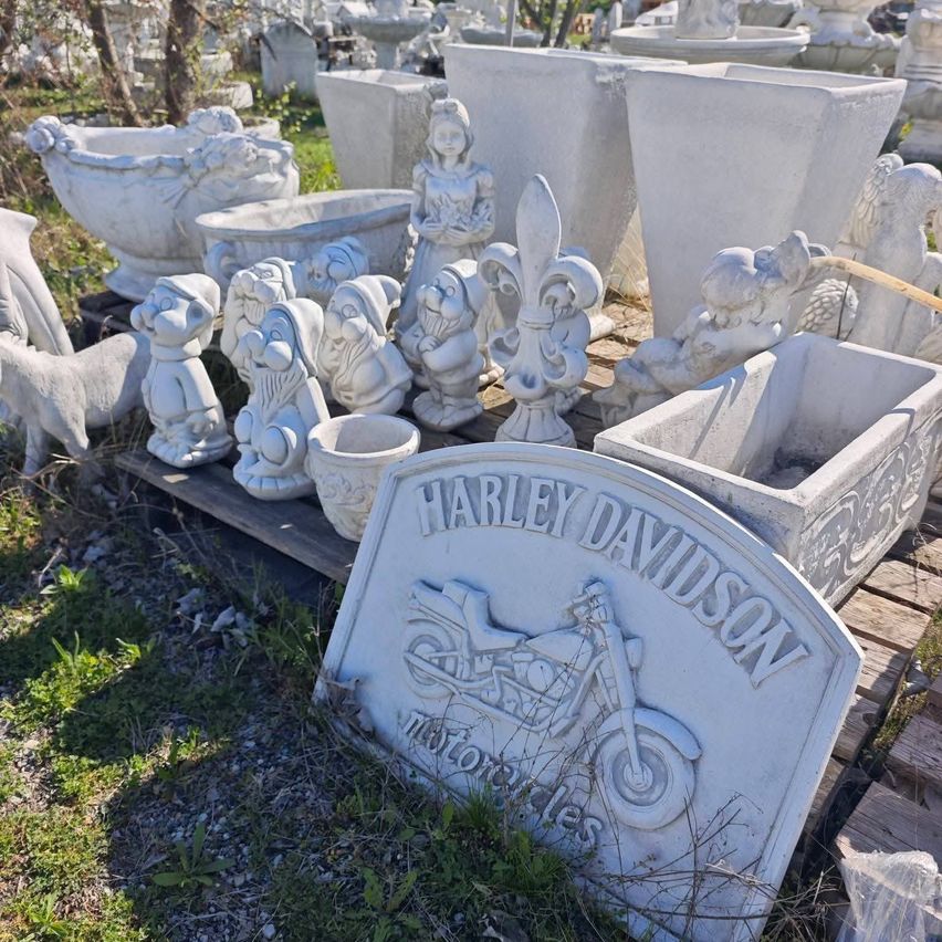 Eine Sammlung von weißen Steingartenfiguren, darunter Gnome, eine Dame und ein Harley-Davidson-Motorradschild, auf einem Holzpalette ausgestellt.