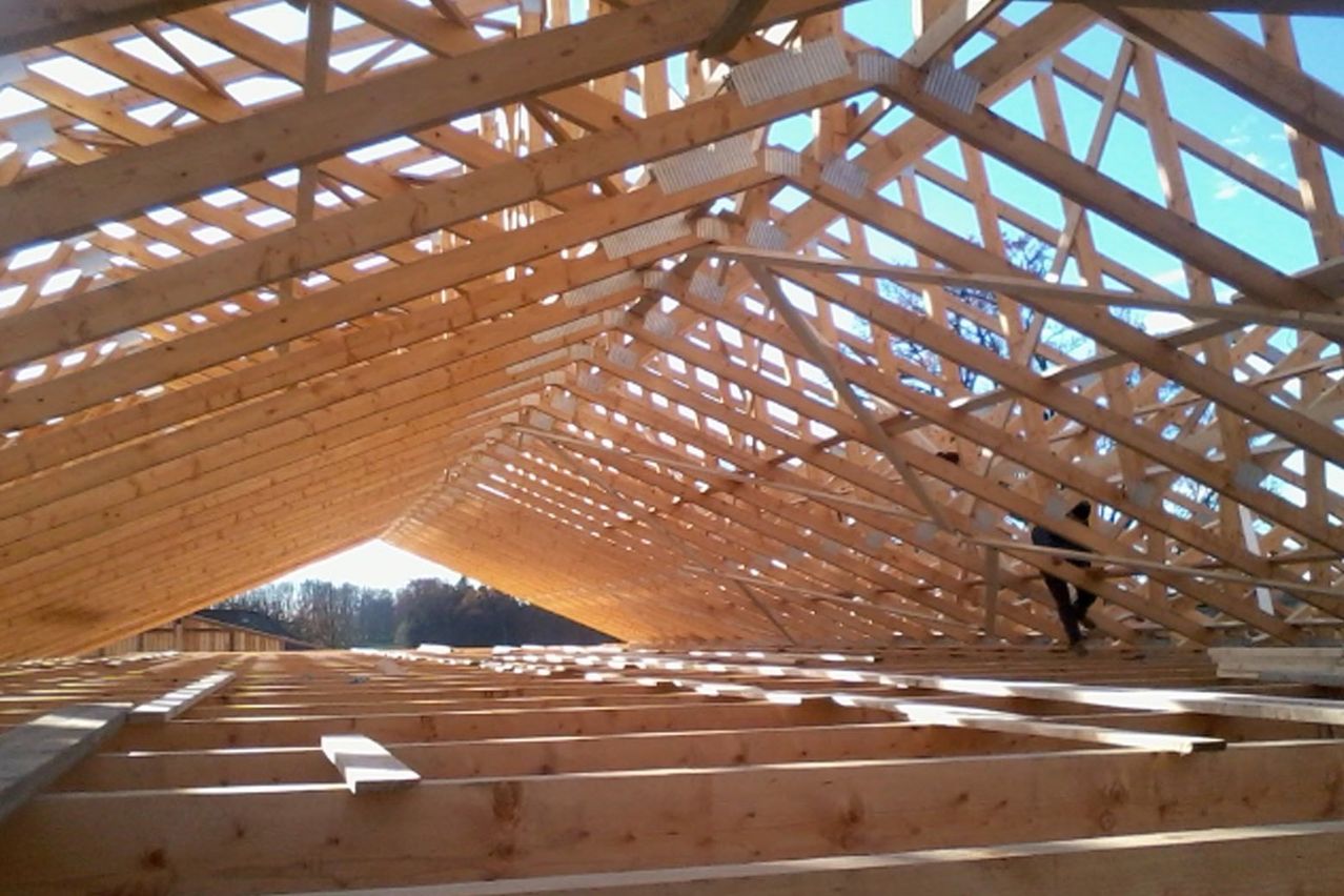 Ein unfertiges Holzdachgerüst mit geometrischem Muster wird errichtet, wobei eine Person auf der rechten Seite steht.