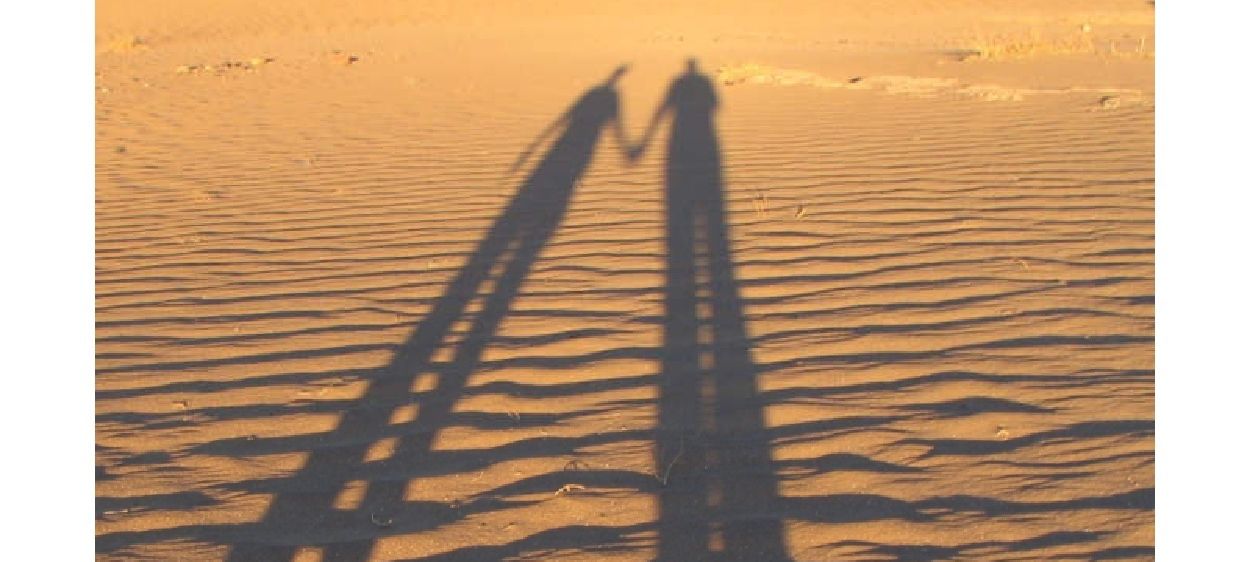 Ein Poster zeigt eine Wüstenszene mit Schatten von zwei Menschen. Es enthält den Text Bleib bitte nicht so, wie du bist! Die Überschrift lautet Volkmarkt - St. Magdalena und März / April 2026.