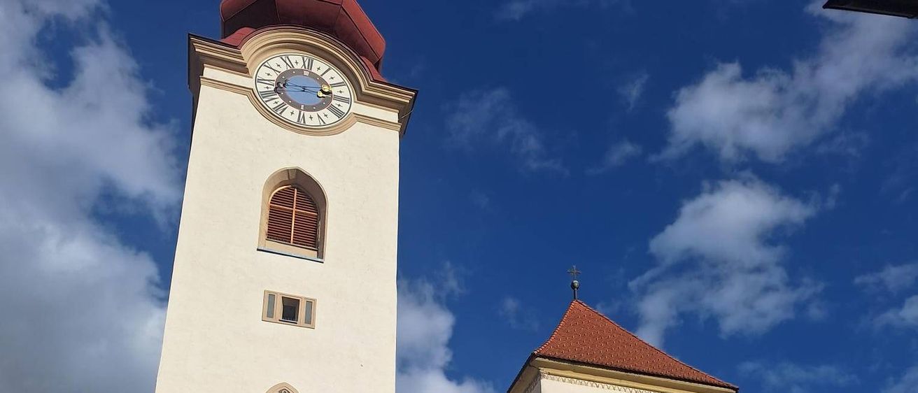 Eine große weiße Kirche mit einem roten Dach und einem hohen Turm, der eine Uhr zeigt. Sie hat bogenförmige Fenster und einen detaillierten Eingang. Das Gebäude steht vor einem blauen Himmel mit verstreuten Wolken.