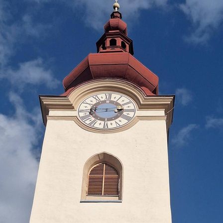 Ein hoher Glockenturm steht gegen einen blauen Himmel mit Wolken. Der Turm hat ein rotes Dach, ein Uhrenblatt und Bogenfenster.