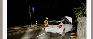 Ein weißer Wagen mit offenem Kofferraum steht am Straßenrand. Ein Verkehrsschild befindet sich am Straßenrand. Ein Feuerwehrmann steht in der Nähe des Autos.