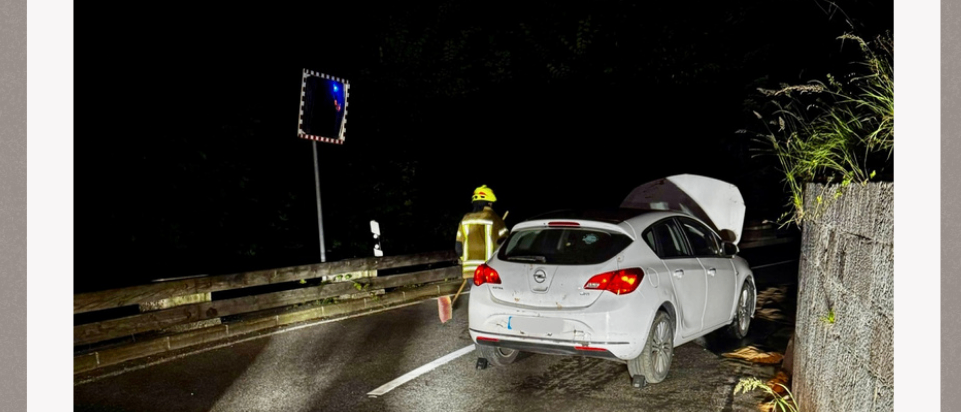 Ein weißer Wagen mit offenem Kofferraum steht am Straßenrand. Ein Verkehrsschild befindet sich am Straßenrand. Ein Feuerwehrmann steht in der Nähe des Autos.