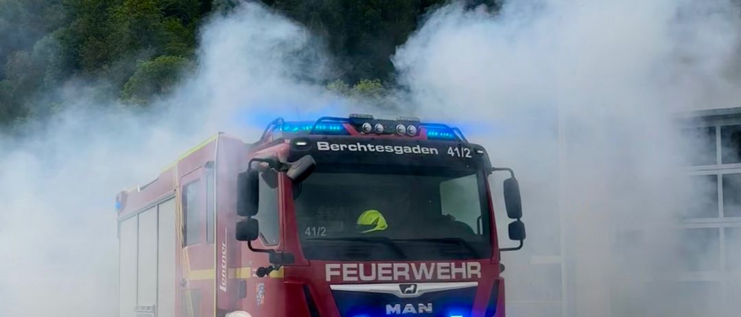 Ein roter Feuerwehrwagen aus Berchtesgaden, Nummer 4112, fährt auf einer Straße und stößt Rauch aus. Dahinter befindet sich ein Gebäude.
