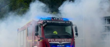 Ein roter Feuerwehrwagen aus Berchtesgaden, Nummer 4112, fährt auf einer Straße und stößt Rauch aus. Dahinter befindet sich ein Gebäude.