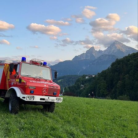 Ein roter Feuerwehrwagen steht auf einer Wiese mit Bergen im Hintergrund. Der Himmel ist klar mit einigen Wolken.