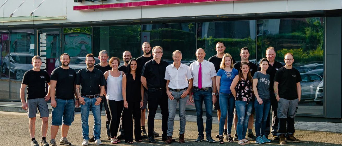 Eine Gruppe von Menschen steht vor dem Auto-Feeberg-Händlergebäude und lächelt für ein Gruppenfoto.
