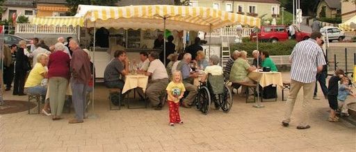 Menschen versammeln sich auf einem Stadtplatz, einige sitzen unter einem gelben und weißen Baldachin an Tischen, andere gehen. Bäume und Gebäude sind im Hintergrund zu sehen, mit einem Kirchturm.