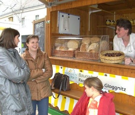 Vier Personen stehen vor einem Bäckereistand, einschließlich einer Frau in einem weißen Shirt. Verschiedene Backwaren sind in Körben ausgestellt. Ein Schild zeigt 'Gloggnitz'.