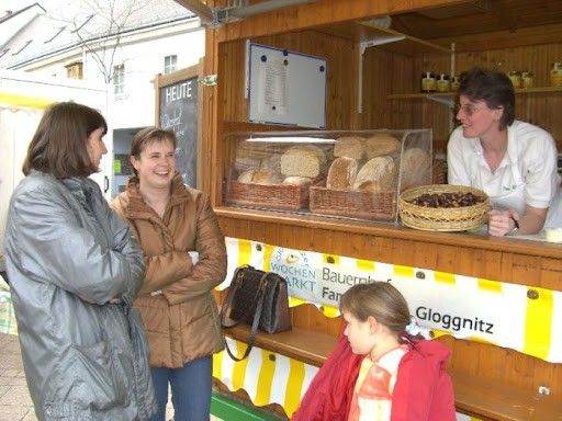 Vier Personen stehen vor einem Bäckereistand, einschließlich einer Frau in einem weißen Shirt. Verschiedene Backwaren sind in Körben ausgestellt. Ein Schild zeigt 'Gloggnitz'.