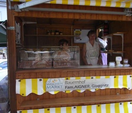 Ein Bäckereistand mit einem gelb-weiß gestreiften Vordach. Zwei Frauen stehen hinter der Theke. Es gibt Regale mit Backwaren und ein Schild, das 'WOCHEN MARKT Bauernhof Fam. AIGNER, Gloggnitz' steht. Ein Auto ist in der Nähe geparkt.