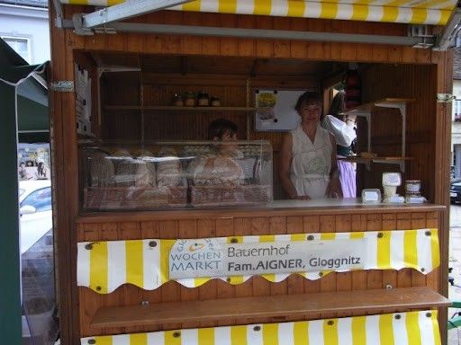 Ein Bäckereistand mit einem gelb-weiß gestreiften Vordach. Zwei Frauen stehen hinter der Theke. Es gibt Regale mit Backwaren und ein Schild, das 'WOCHEN MARKT Bauernhof Fam. AIGNER, Gloggnitz' steht. Ein Auto ist in der Nähe geparkt.