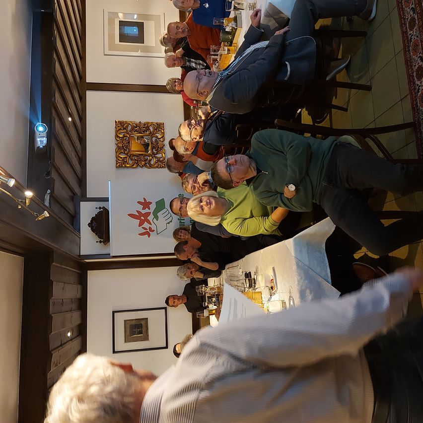 Eine Gruppe von Menschen sitzt an einem langen Tisch in einem Restaurant. Einige tragen Brillen, einer hat eine Uhr. Es gibt Bilderrahmen an der Wand und ein Banner mit einem Logo.
