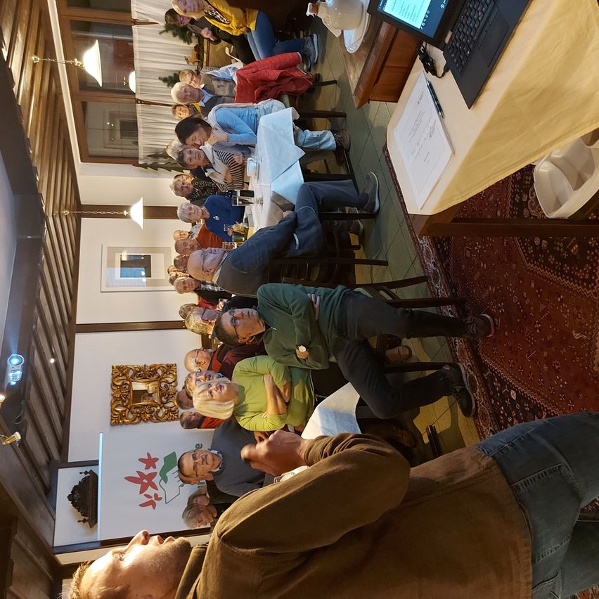 Eine Gruppe von Menschen sitzt in einem Raum, wahrscheinlich bei einer Besprechung. Ein Mann steht vorne, andere lauschen. Tische und Stühle sind mit einem Laptop und Papieren ausgestattet.