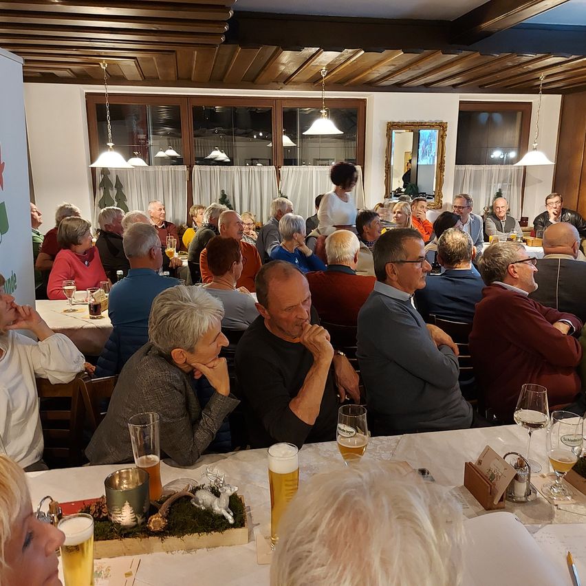 Ein großer Speisebereich mit vielen Menschen, einige sitzen an Tischen mit Weingläsern und Bierkrügen. Eine Frau steht in der Mitte, und andere führen Gespräche. Der Raum hat Holzdecken, hängende Lampen und Fenster mit Vorhängen.