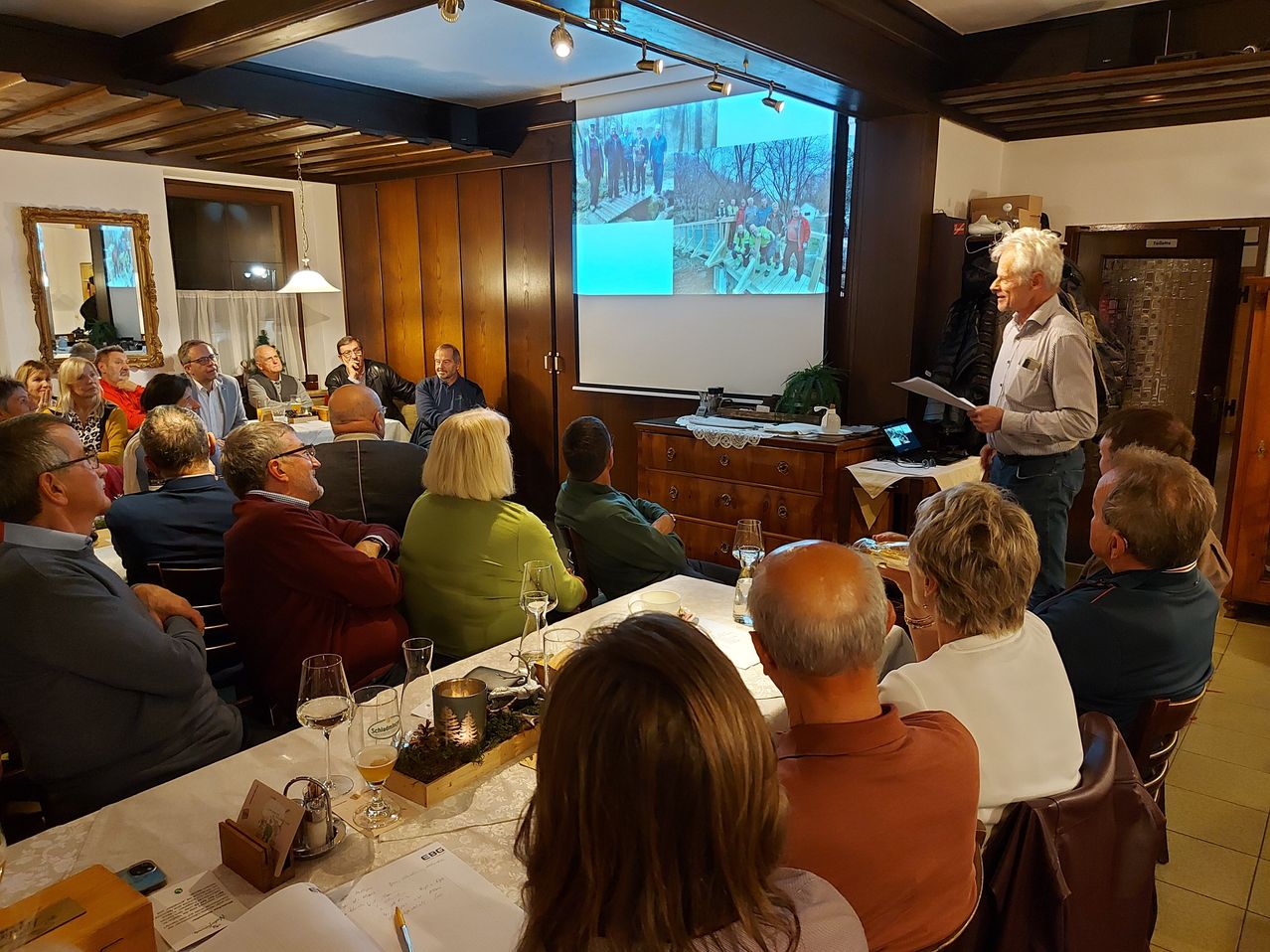 Eine Gruppe von Menschen versammelt sich in einem Raum und schaut sich eine Präsentation auf einer Projektionsleinwand an. Der Präsentator steht vor einem Tisch mit einer Pflanze und einem Laptop. Die Teilnehmer sitzen um einen Tisch mit Weingläsern und einer Speisekarte. Der Raum hat Holzwände und eine Decke mit Beleuchtung.