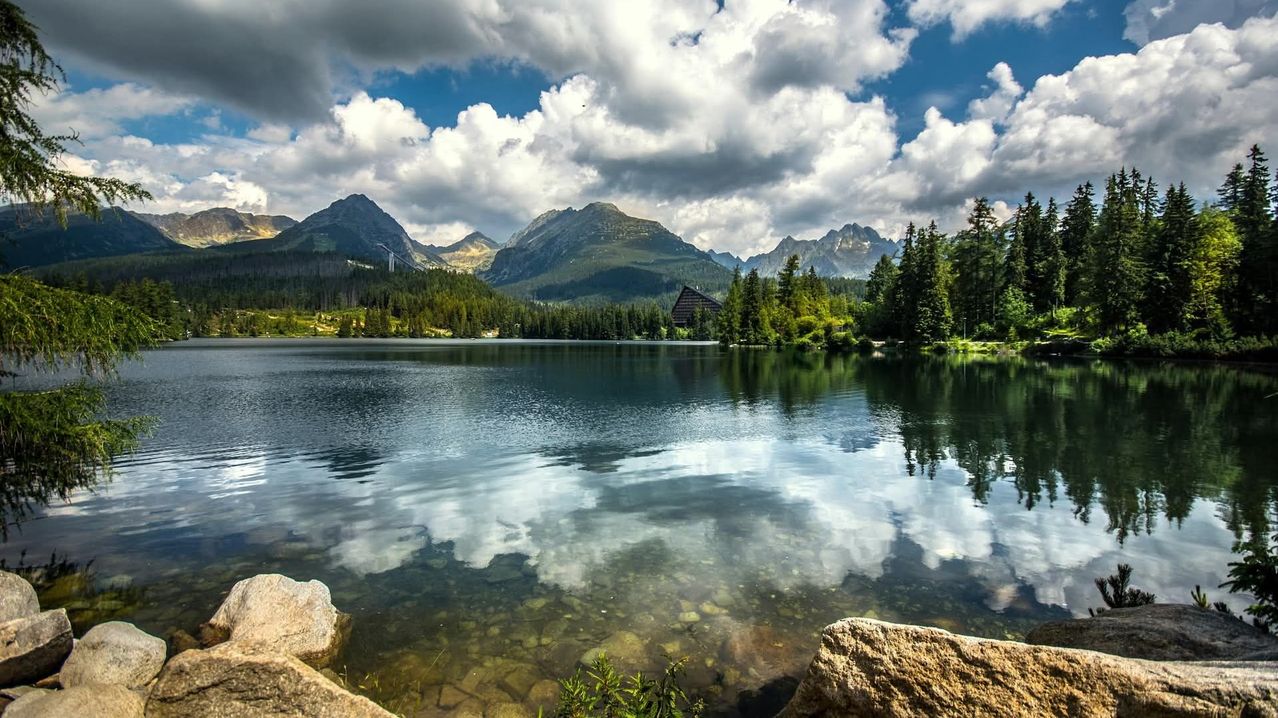 A serene lake with rocky shoreline, reflecting the sky and clouds, surrounded by lush green trees and mountains in the distance.