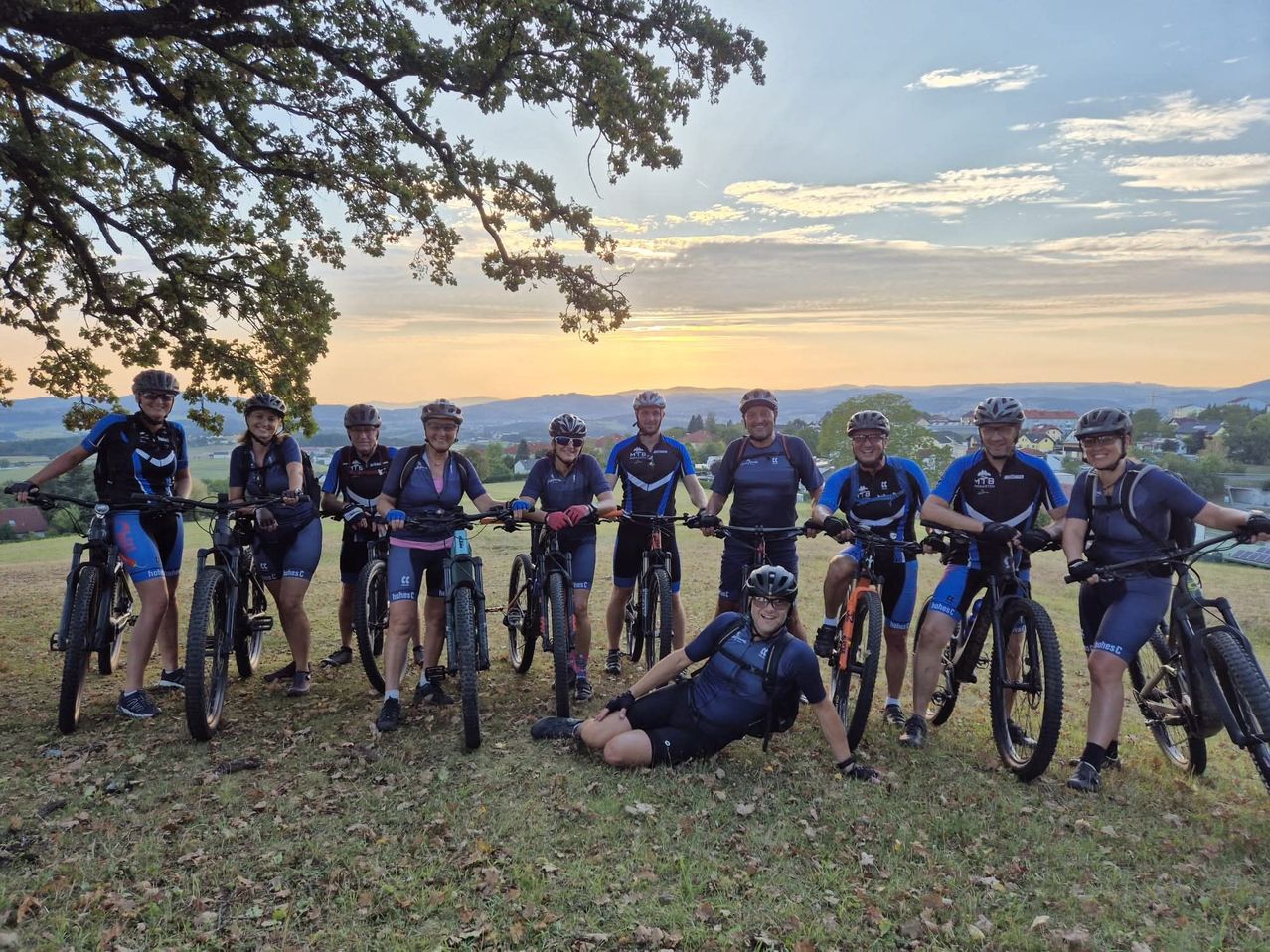Eine Gruppe von Radfahrern, einige sitzend und andere stehend, posiert für ein Foto bei Sonnenuntergang mit Helmen, Fahrrädern in der Hand und einer malerischen Aussicht.