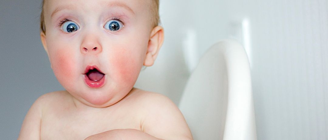 Ein Baby mit blauen Augen sitzt in einem Hochstuhl, der Mund ist offen und es schaut nach oben. Die Hände des Babys liegen auf dem Tisch, und seine Wangen sind leicht gerötet.