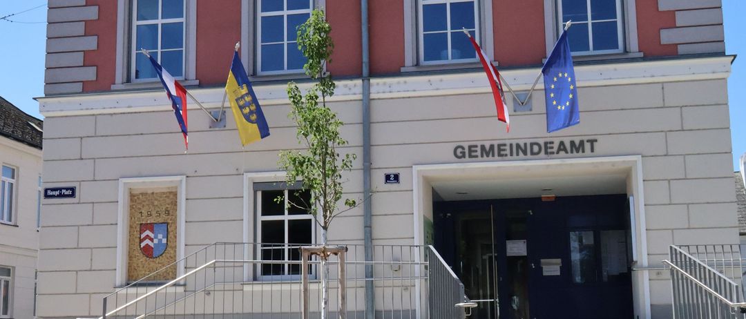 Das Gemeindeamtsgebäude hat eine rote Fassade mit weißen Umrandungen. Es wehen zwei Fahnen, eine davon ist die EU-Fahne. Der Eingang hat eine Treppe, die zu ihm führt.