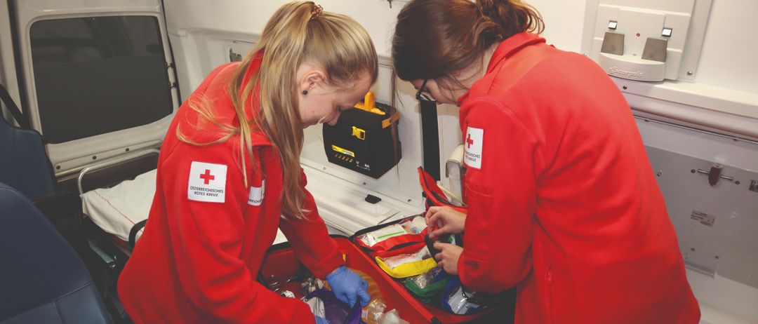 Zwei Frauen in roten Uniformen mit rotem Kreuzsymbol arbeiten in einem Fahrzeug, möglicherweise einem Krankenwagen. Sie bereiten wahrscheinlich medizinische Ausrüstung vor.