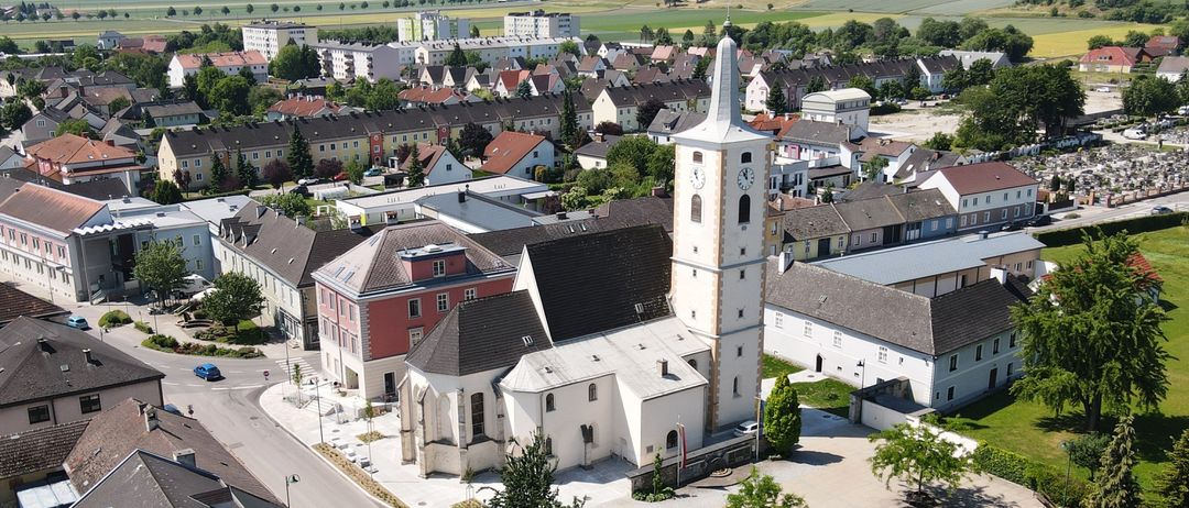 Luftaufnahme einer Stadt mit einer Kirche mit hohem Turm, umgeben von Gebäuden, Bäumen und einer Straße. Die Landschaft umfasst auch ländliche Gebiete und Berge in der Ferne.