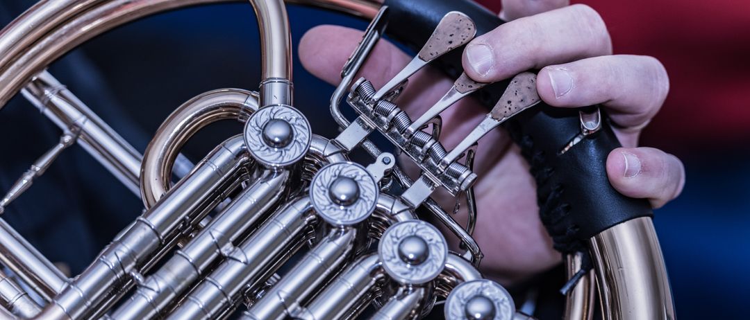 Nahaufnahme von Händen, die die Ventile einer silbernen Waldhorns einstellen. Das komplizierte Design und der polierte Glanz des Instruments werden hervorgehoben.