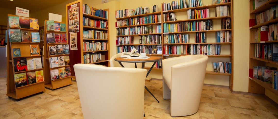 Ein Innenansicht einer kleinen Bibliothek mit gelber Wand und gefliestem Boden. Zwei weiße Stühle umgeben einen kleinen Tisch mit offenem Buch. Regale mit Büchern dahinter.