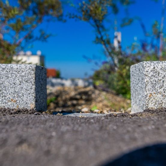Eine Nahaufnahme von zwei großen Granitblöcken auf einer Straße mit Grünpflanzen im Hintergrund unter einem blauen Himmel.