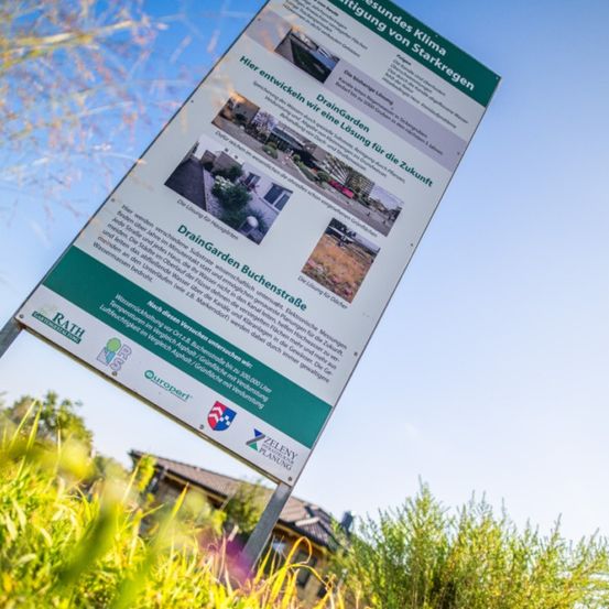 Ein Schild mit dem Titel 'DrainGarden' handelt von nachhaltiger Wasserwirtschaft. Es steht in einem Grasbereich. Es hat Bilder und Text, von denen einige auf Deutsch sind.