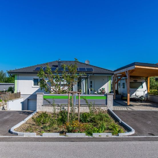 Ein modernes Haus mit Solarpanelen auf dem Dach, einem kleinen Garten davor und einem Auto, das unter einem Holzcarport geparkt ist.