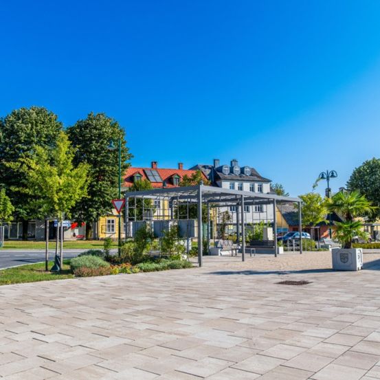Ein sonniger Park mit einem Metallpavillon, umgeben von Bäumen und Pflanzen. In der Ferne befinden sich Gebäude, eines mit Solarpanelen auf dem Dach.