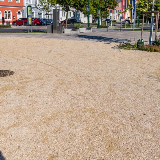 Ein breiter, leerer Kiesbereich in einer städtischen Außenumgebung. Bäume und Gebäude sind im Hintergrund. Eine Person geht in der Nähe der Gebäude.