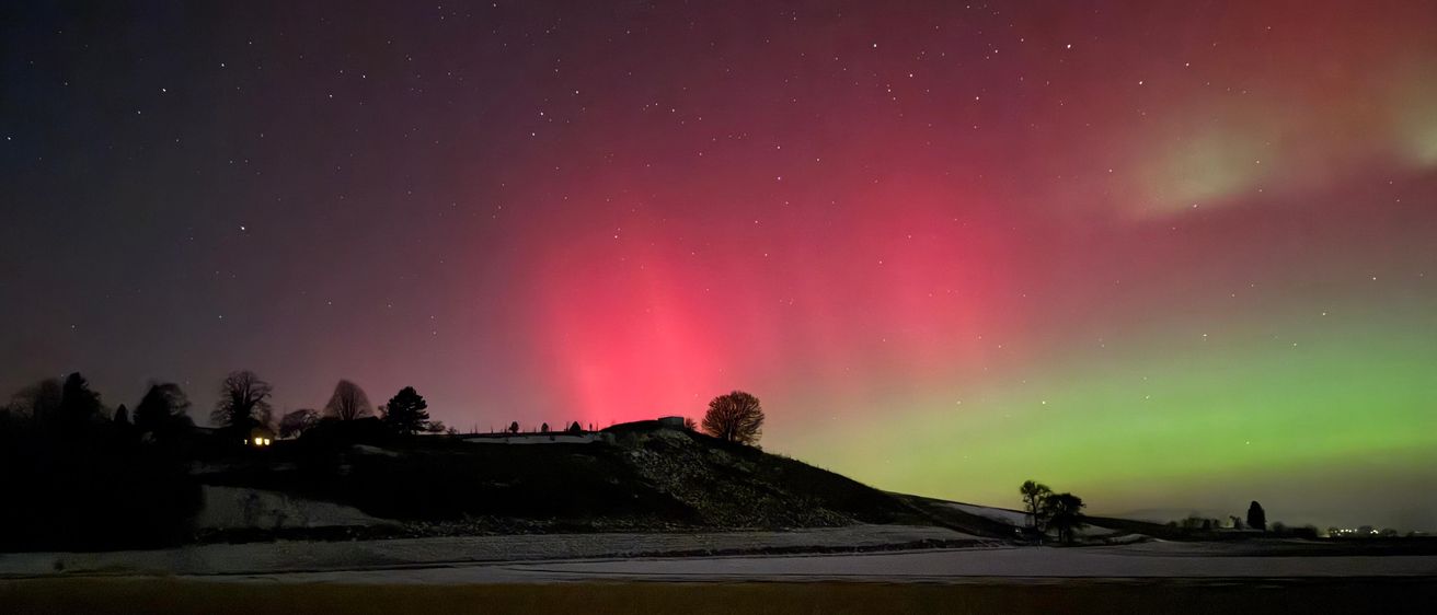 Ein Nachthimmel, der vom Polarlicht erleuchtet wird, mit leuchtenden roten und grünen Lichtern, die sich über den Himmel erstrecken. Bäume und ein Gebäude sind im Vordergrund sichtbar, und die Landschaft ist mit Schnee bedeckt.