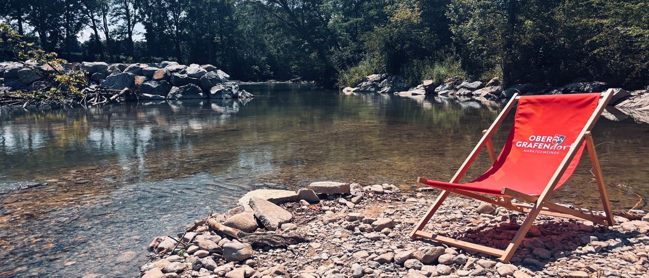 Ein ruhiger Fluss mit felsigen Ufern und einem roten Liegestuhl am Ufer. Das Wasser ist klar und reflektiert die grünen Bäume.