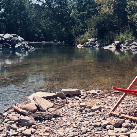 Ein ruhiger Fluss fließt durch ein bewaldetes Gebiet. Felsige Ufer säumen das Wasser, und ein roter Stuhl steht am Ufer.