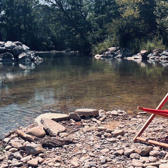 Ein ruhiger Fluss fließt durch ein bewaldetes Gebiet. Felsige Ufer säumen das Wasser, und ein roter Stuhl steht am Ufer.