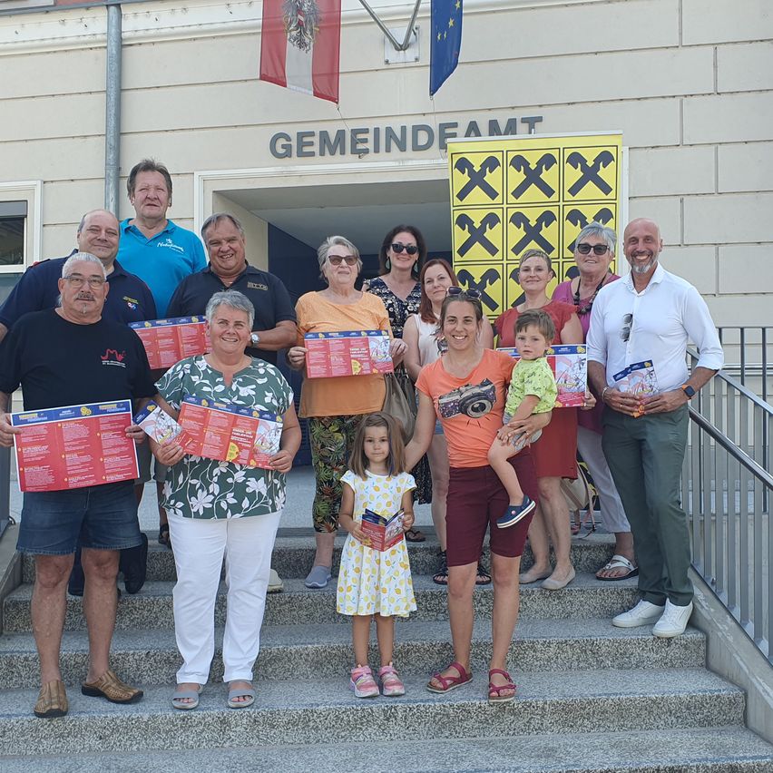 Eine Gruppe von Menschen, darunter Erwachsene und Kinder, posiert für ein Foto auf den Stufen eines Gebäudes, hält Broschüren und lächelt. Hinter ihnen befindet sich ein gelbes Schild mit einem Schachbrettmuster.