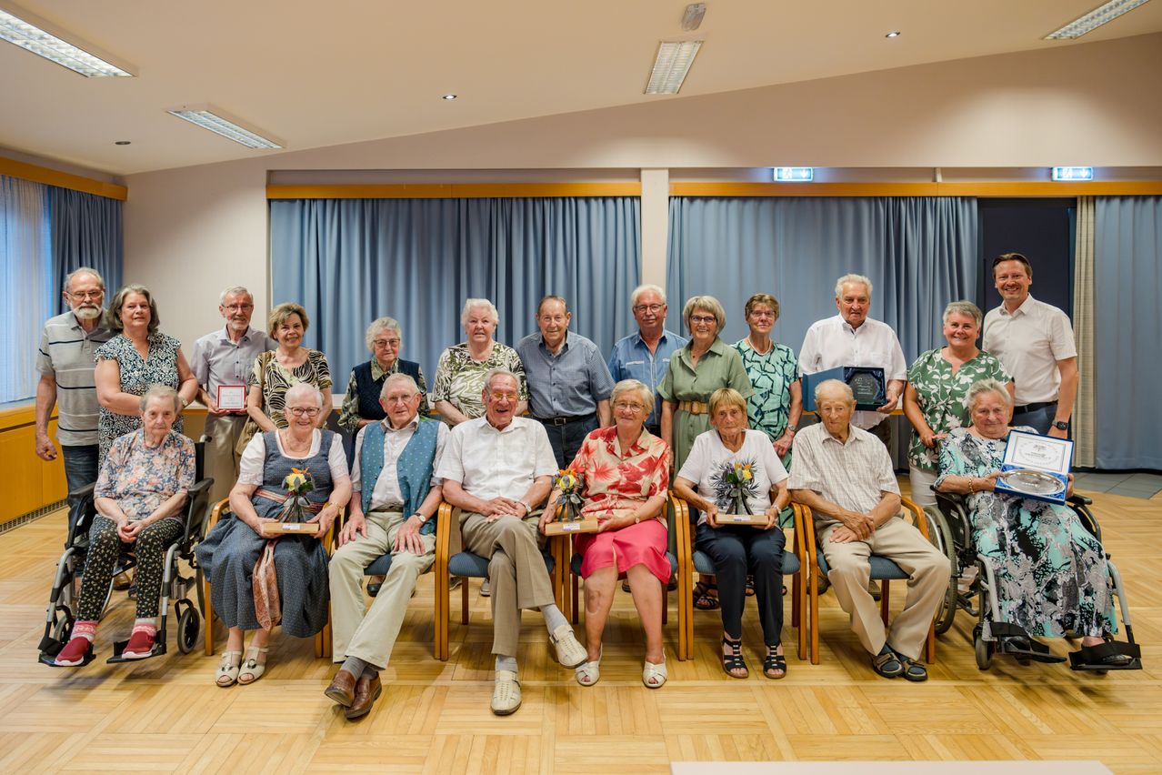 Eine Gruppe älterer Menschen versammelt sich in einem Raum. Sie sitzen auf Stühlen und halten Auszeichnungen. Sie tragen formelle Kleidung und einige von ihnen tragen Brillen. Sie posieren für ein Foto. Der Raum hat blaue Vorhänge und hölzerne Böden.