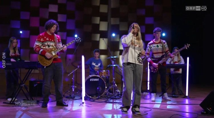 Eine Gruppe junger Musiker tritt auf der Bühne auf. Ein Mann spielt Gitarre, während eine Frau singt und die Schellentrommel spielt. Dahinter spielen zwei andere Musiker Schlagzeug und Keyboard. Die Bühne ist mit lila und blauen Lichtern beleuchtet.