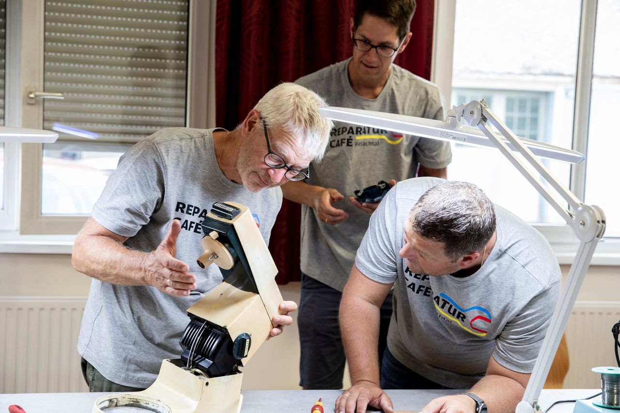 Drei Männer arbeiten an einer Maschine, einer von ihnen zeigt darauf. Sie tragen graue T-Shirts. Auf einem steht 'Reparatur Cafe'. Ein anderer Mann schaut über die Schulter.