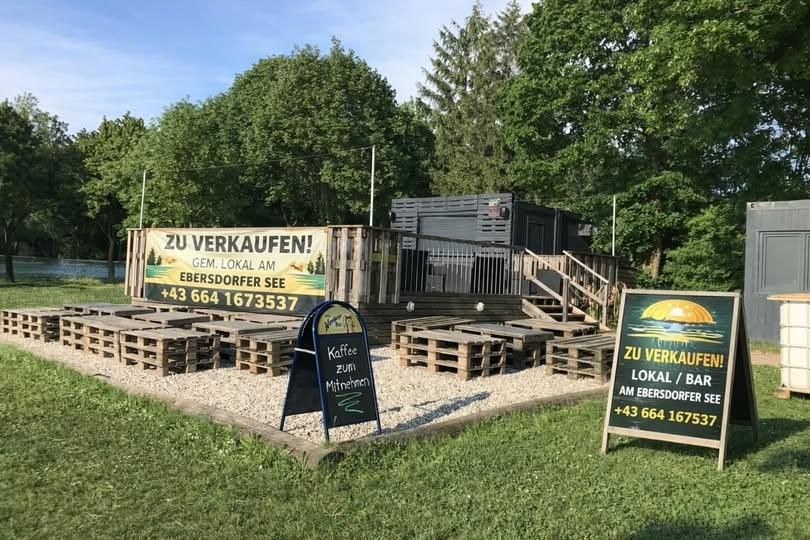 Ein Holzcafé am Ebersdorfer See mit Außenbestuhlung, einer Treppe und einem Schild, das Kaffee zum Mitnehmen anbietet. Ein großes Banner bewirbt den Verkauf des Cafés.