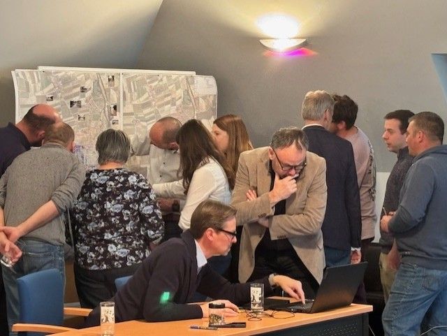 Mehrere Personen versammeln sich in einem Raum, wobei ein Mann einen Laptop an einem Schreibtisch benutzt. Der Raum ist hell beleuchtet, und eine Karte hängt an der Wand.