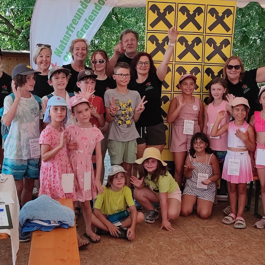 Eine Gruppe von Kindern und Erwachsenen posiert für ein Foto vor einem gelben Banner mit schwarzen Xs. Einige tragen Hüte und Brillen.